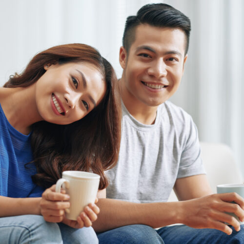 happy-asian-couple-sitting-couch-home-with-tea-mugs-smiling