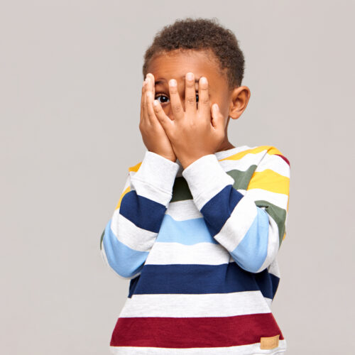 Frightened dark skinned little boy covering face with both hands as if being afraid to see something scary, spying through hole between fingers. Shy African child hiding, or playing hide and seek